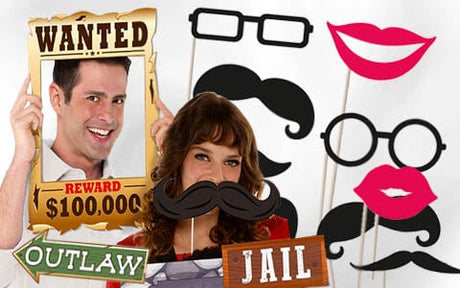 Zwei Personen posieren vor einem weißen Hintergrund mit Requisiten aus einer Fotokabine, darunter ein Steckbrief, Schnurrbärte, Brillen, Lippen sowie Schilder mit den Aufschriften „OUTLAW“ und „JAIL“.