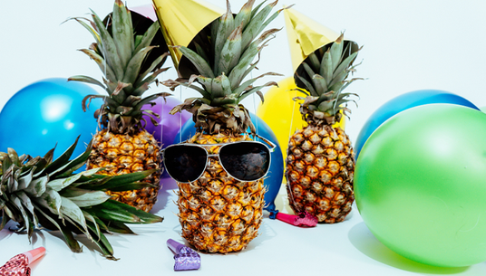 Drei Ananas mit Partyhütchen inmitten bunter Luftballons; eine Ananas trägt eine Sonnenbrille.