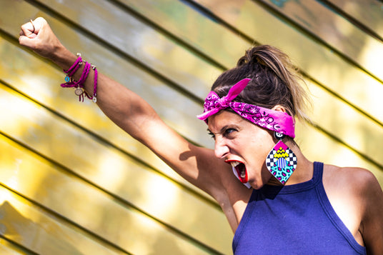 Eine Frau in einem blauen Tanktop und einem rosa Bandana steht vor einer gelb gestreiften Wand, reckt die Faust in die Höhe und ruft laut.