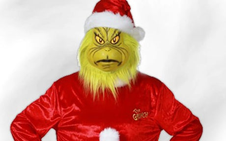 Eine Person in einem Grinch-Kostüm mit roter Weihnachtsmannmütze und rotem Anzug, die mit den Händen in den Hüften vor einem schlichten weißen Hintergrund steht.