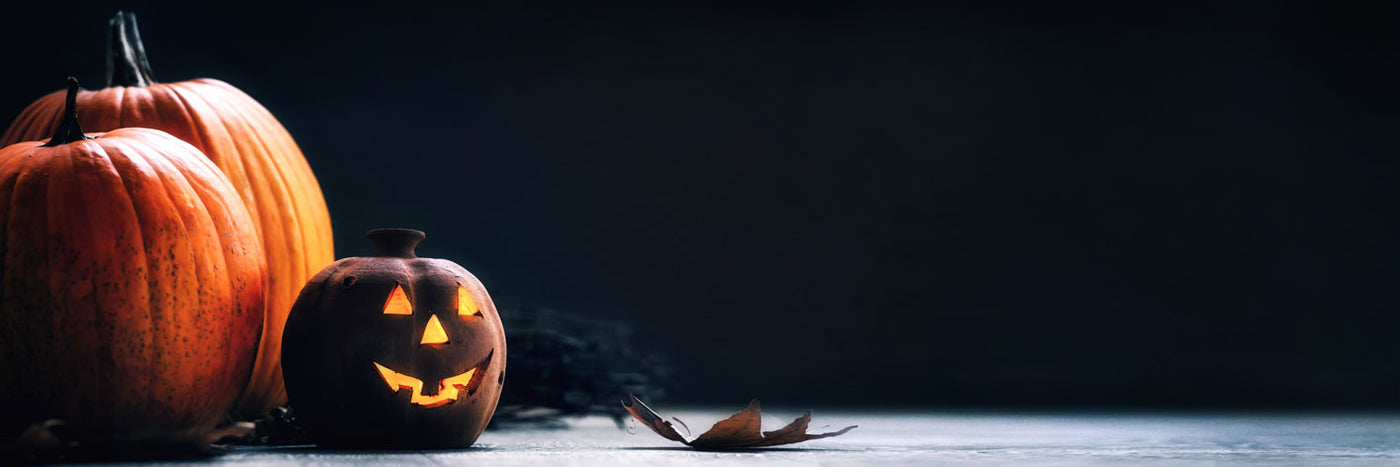 Drei Kürbisse, von denen einer als Jack-o-Lantern mit leuchtendem Gesicht geschnitzt ist, stehen auf einer dunklen Fläche neben einem trockenen Blatt und bei gedämpfter Beleuchtung.