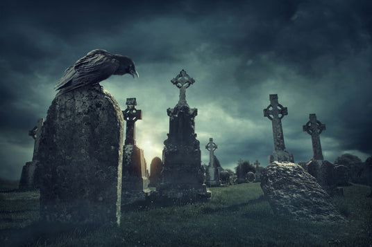 Ein Rabe sitzt auf einem verwitterten Grabstein auf einem alten Friedhof mit keltischen Kreuzen unter einem dunklen, bewölkten Himmel.