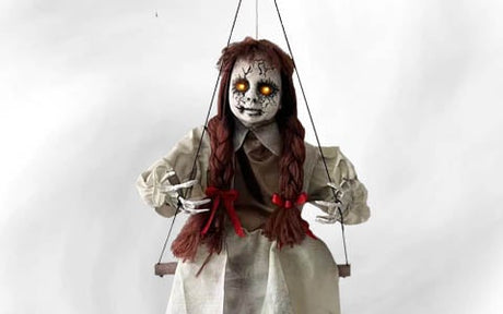 Eine Marionette mit leuchtend gelben Augen, einem rissigen Gesicht, langen braunen Zöpfen und einem weißen Kleid, die an Fäden vor einem schlichten Hintergrund hängt.