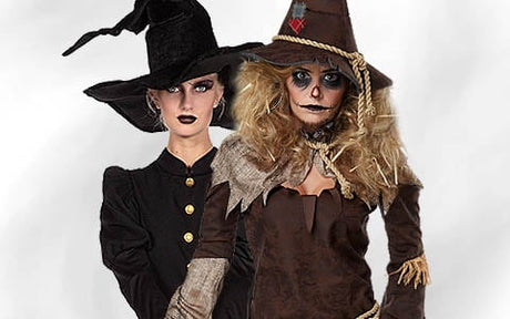 Zwei Personen in Halloween-Kostümen: Die eine ist als Hexe in Schwarz mit spitzem Hut verkleidet, die andere als Vogelscheuche mit braunem Hut, Details aus Sackleinen und einer Gesichtsbemalung, die an Nähte erinnert.