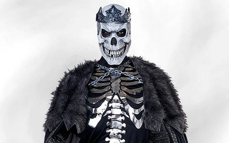 Eine Person in einem Skelettkostüm mit Krone und pelzbesetztem Umhang, mit Kettenverzierungen und einer Totenkopfmaske, die vor einem schlichten, hellen Hintergrund steht.