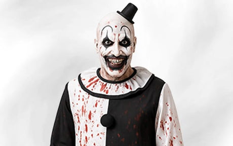 Eine Person, verkleidet als gruseliger Clown mit schwarz-weißem Make-up, einem kleinen Hut und einem blutbefleckten Clownskostüm, lächelt breit vor einem schlichten Hintergrund.
