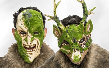 Zwei Personen tragen grün-weiße Masken mit moosartiger Struktur; eine Maske sieht menschenähnlich aus und knurrt, die andere ähnelt einem Reh mit Geweih. Beide tragen Kleidung aus braunem Fell.