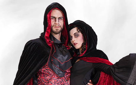 Ein Mann und eine Frau tragen passende schwarz-rote Vampir-Kostüme mit Kapuzen, auffälligem Make-up und ernsten Gesichtsausdrücken und posieren vor einem schlichten weißen Hintergrund.