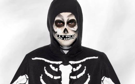Eine Person in einem schwarzen Skelettkostüm mit Kapuze, das mit weißen Knochenmustern verziert ist, und mit Totenkopf-Gesichtsbemalung, die vor einem hellen Hintergrund direkt in die Kamera blickt.