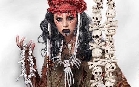 Eine Person in Kostüm mit schwarzen Haaren, rotem Kopfschmuck, Gesichtsbemalung und Accessoires aus Knochen und Schädeln, die vor einem schlichten Hintergrund steht.