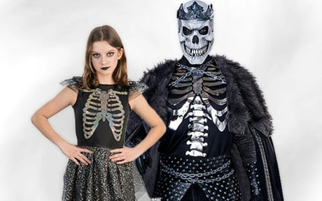 Ein Mädchen in einem Brustkorbkostüm steht neben einer Person in einem Skelettkönig-Kostüm mit Totenkopfmaske, Krone und schwarzem Umhang.