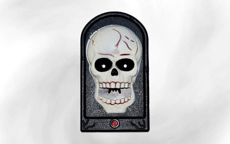 Auf einer grabsteinartigen Plakette, an deren Unterseite sich ein roter Knopf befindet, ist ein karikaturhafter Totenkopf mit schwarzen Augen und einer rissigen Stirn abgebildet.