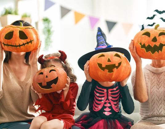 Vier Personen in Halloween-Kostümen sitzen in einem Raum und halten sich geschnitzte Kürbisse vor das Gesicht. Im Hintergrund hängen bunte Wimpelketten.