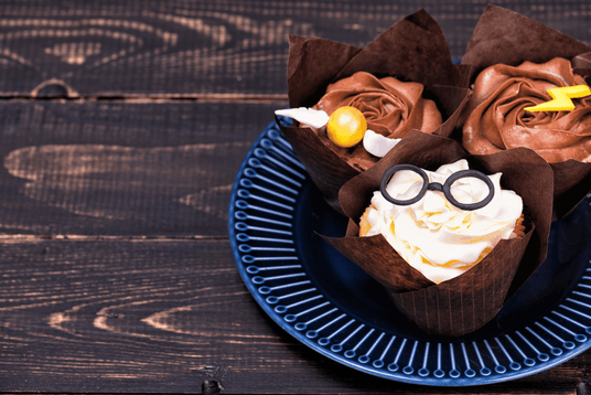 Drei Cupcakes auf einem blauen Teller, zwei mit Schokoladenglasur und einer mit weißer Glasur, verziert mit bonbonförmigen Brillen, runden Bonbons und einem gelben Blitzbonbon.