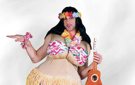 Eine Person in einem Hula-Kostüm mit einem Blumenkranz, einem Grasrock, einer schwarzen Perücke und Blumenaccessoires, die eine kleine Ukulele vor einem schlichten Hintergrund in der Hand hält.