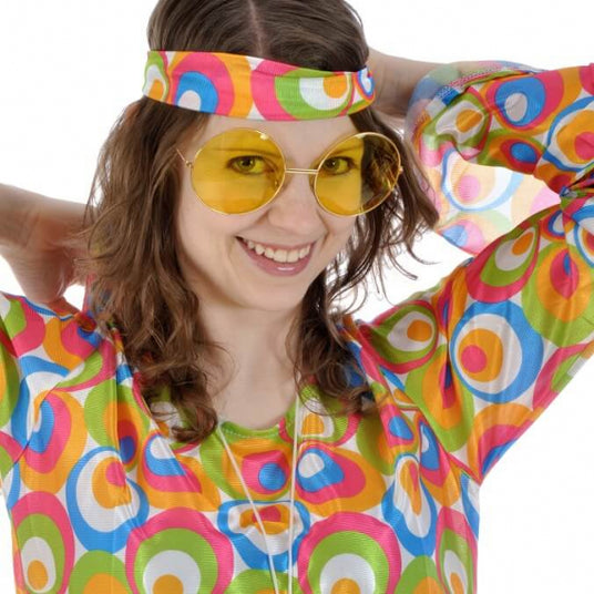 Eine Person, die ein buntes Oberteil mit Retro-Muster, eine gelb getönte runde Sonnenbrille und ein dazu passendes Stirnband trägt und mit hinter dem Kopf verschränkten Händen lächelt.