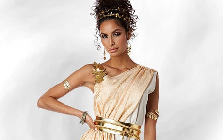 Eine Frau in einem gold- und cremefarbenen, an eine Toga angelehnten Kleid mit goldenen Accessoires – darunter ein Kopfschmuck, Ohrringe, ein Armband und Gürtel –, posiert vor einem hellen Hintergrund.