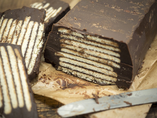 Ein in Scheiben geschnittener Schokoladen-Keks-Kuchen mit sichtbaren Schichten aus Schokolade und Keksen, der auf braunem Backpapier liegt, daneben liegt ein Messer.