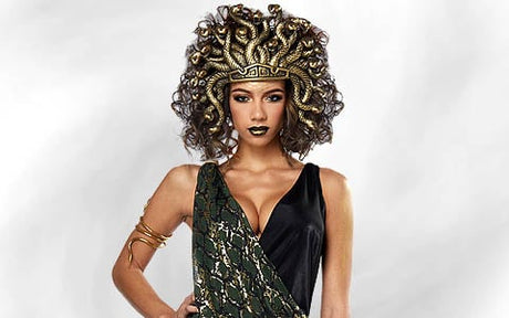 Eine Frau in einem Medusa-Kostüm mit einem Schlangenkopfschmuck, dunklem Lippenstift, einem einseitigen Kleid und goldenem Armschmuck, die vor einem hellen Hintergrund steht.