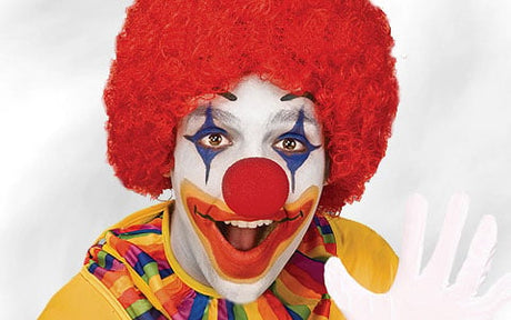 Eine Person, die als Clown verkleidet ist – mit roter Perücke, weißer Gesichtsbemalung, buntem Kostüm und einer großen roten Nase –, lächelt und winkt in die Kamera.