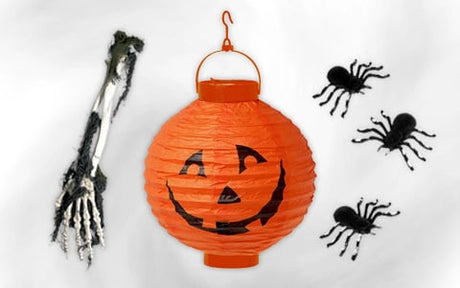 Auf einem weißen Hintergrund sind eine orangefarbene Papierlaterne mit einem Kürbiskopf, eine Skelett-Hand und drei schwarze Spinnen-Verzierungen angeordnet.