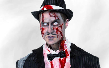 Eine Person in einem Nadelstreifenanzug und mit Hut, mit Zombie-Make-up, weißen Kontaktlinsen und Kunstblut im Gesicht und auf der Kleidung, vor einem schlichten Hintergrund.
