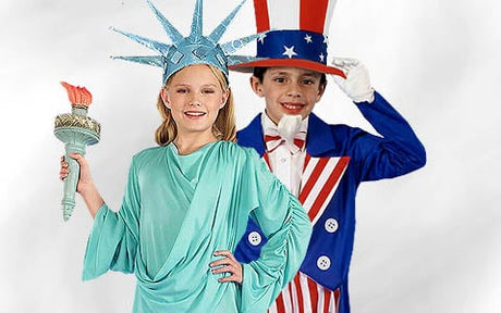 Zwei Kinder in Kostümen: Das eine ist als Freiheitsstatue verkleidet und hält eine Fackel in der Hand, das andere als Uncle Sam, der seinen Hut lüftet; beide stehen nebeneinander vor einem schlichten Hintergrund.