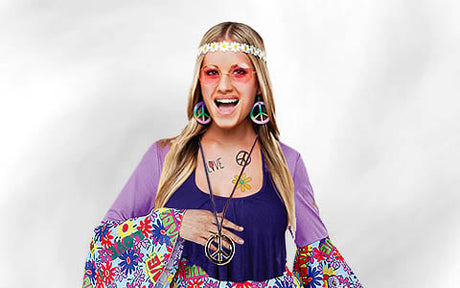 Eine Person in einem farbenfrohen Hippie-Kostüm im Stil der 1970er Jahre mit Accessoires mit Peace-Zeichen, Blumenmustern, einem Stirnband und einer runden Sonnenbrille, die vor einem hellen Hintergrund lächelt.