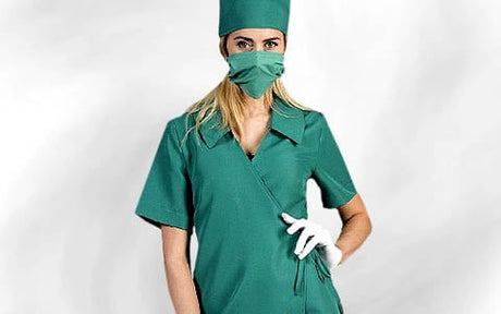 Person in grüner OP-Kleidung, mit Haube, Maske und weißen Handschuhen, vor einem schlichten, hellen Hintergrund stehend.