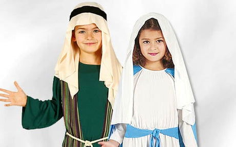 Zwei Kinder in traditionellen Krippenkostümen, eines als Josef in Grün und das andere als Maria in Weiß und Blau, stehen vor einem schlichten weißen Hintergrund.