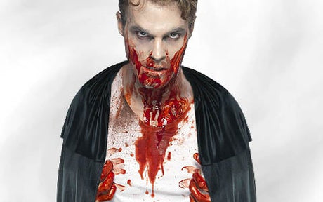Ein Mann mit blasser Haut und dunklem Make-up, der einen schwarzen Umhang und ein weißes Hemd trägt, hat Kunstblut an Mund, Händen und Brust und steht vor einem schlichten weißen Hintergrund.