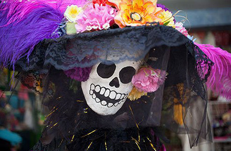 Unter einem großen schwarzen Hut, der mit bunten Blumen und violetten Federn verziert ist, lächelt eine bemalte Totenkopfmaske mit großen Zähnen.