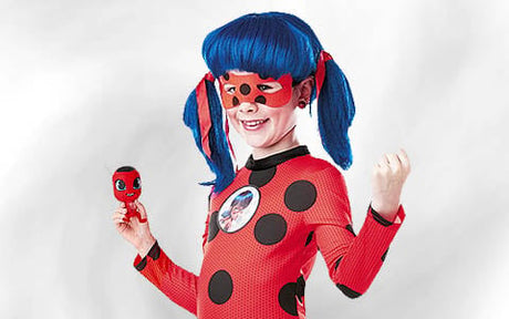 Ein Kind in einem rot-schwarz gepunkteten Kostüm mit einer blauen Zopfperücke und einer roten Maske, das eine kleine, dazu passende Puppe in der Hand hält und vor einem schlichten weißen Hintergrund posiert.
