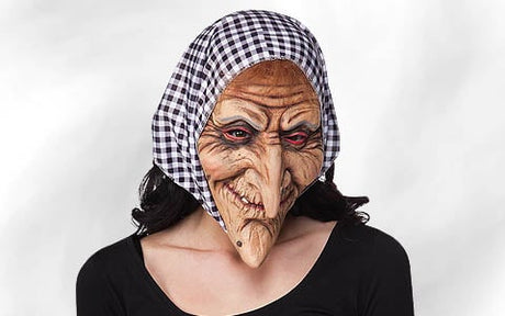 Eine Person, die eine zerknitterte Hexenmaske mit langer Nase und langem Kinn sowie roten Augen trägt und ein schwarz-weiß kariertes Kopftuch auf dem Kopf hat, vor einem einfarbigen Hintergrund.