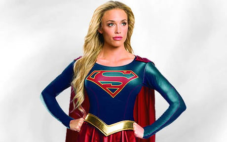 Eine Person mit langen blonden Haaren, verkleidet als Supergirl, steht mit den Händen in den Hüften vor einem schlichten Hintergrund und trägt ein blau-rotes Kostüm mit einem roten S-Emblem und einem roten Umhang.