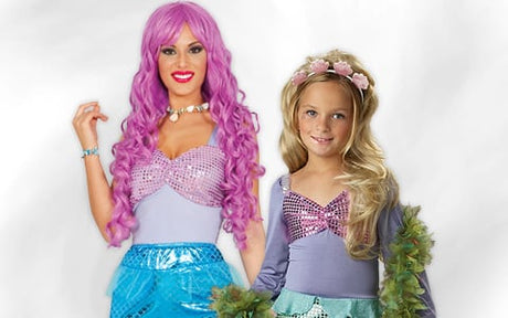 Zwei Personen in Meerjungfrauenkostümen – ein Erwachsener mit langen rosa Haaren und ein Kind mit langen blonden Haaren –, beide mit schimmernden Oberteilen und blauen Schwänzen, stehen vor einem hellen Hintergrund.