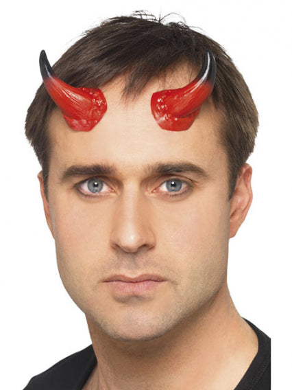 Ein Mann mit kurzen braunen Haaren und blauen Augen trägt rote und schwarze Teufelshörner als Accessoire auf der Stirn und blickt direkt in die Kamera.