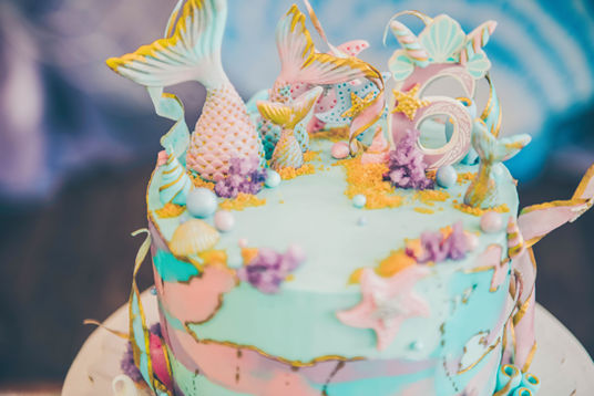 Bunter Kuchen im Meerjungfrauen-Stil mit Schwänzen, Muscheln und Meeresdekorationen, auf dessen Oberseite die Zahl sechs zu sehen ist.