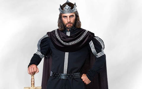 Ein Mann, gekleidet wie ein mittelalterlicher König, mit einer Krone und einem dunklen Gewand, der ein Schwert hält, wobei eine Hand auf dem Griff ruht, und vor einem schlichten, hellen Hintergrund steht.
