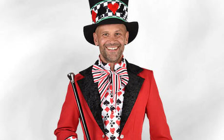 Ein Mann mit einem hohen Hut und einem roten Anzug, der mit Spielkartensymbolen verziert ist, hält einen Spazierstock in der Hand und lächelt in die Kamera.