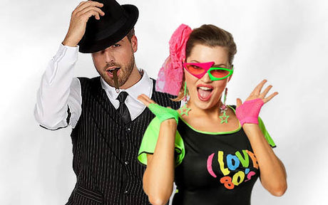 Ein Mann in einem Nadelstreifenanzug und einem Fedora steht neben einer Frau in bunter 80er-Jahre-Kleidung mit Neon-Accessoires und einem „Love 80s“-T-Shirt.