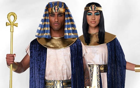Zwei Personen in Kostümen im Stil des alten Ägyptens mit goldenen Accessoires und blau-goldenen Kopfbedeckungen, die nebeneinander vor einem schlichten Hintergrund stehen.
