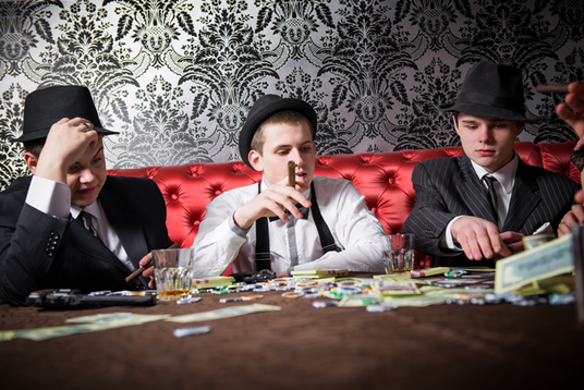 Drei junge Männer in Anzügen und mit Hüten sitzen an einem Tisch mit Spielkarten, Pokerchips, Bargeld, Zigarren und Getränken in einem schwach beleuchteten Raum im Vintage-Stil.