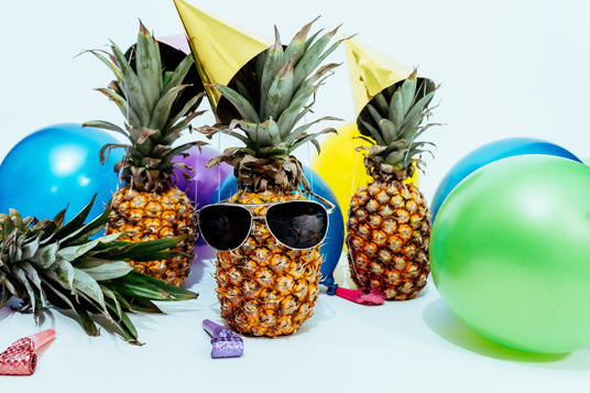 Drei Ananas mit Partyhütchen, umgeben von bunten Luftballons; die vordere Ananas trägt eine Sonnenbrille.