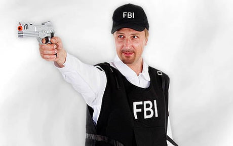 Eine Person in einer FBI-Weste und einer FBI-Mütze richtet eine Spielzeugpistole mit orangefarbener Spitze nach vorne; der Hintergrund ist einfarbig weiß.