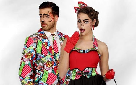 Zwei Personen tragen farbenfrohe Kostüme im Pop-Art-Stil und auffälliges Make-up. Der Mann trägt einen Anzug und eine Krawatte im Comic-Stil, während die Frau ein rotes Kleid und Handschuhe trägt; beide posieren vor einem schlichten Hintergrund.