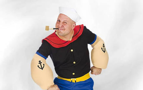 Eine Person in einem Cartoon-Matrosenkostüm mit einem weißen Hut, übergroßen, gepolsterten Ärmeln mit Anker-Tattoos, einem rot-schwarzen Hemd, einer blauen Hose und einer Pfeife im Mund.