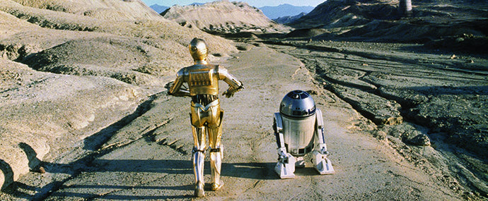Zwei Roboter – ein goldfarbener, humanoider und ein kleiner, zylindrischer – gehen unter klarem Himmel Seite an Seite durch eine karge, felsige Wüstenlandschaft.
