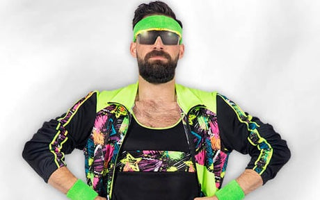 Ein Mann mit einem neongrünen Stirnband, einer Sonnenbrille und einem bunten Trainingsoutfit im 80er-Jahre-Stil mit passenden Armstulpen, der mit den Händen in den Hüften vor einem schlichten weißen Hintergrund steht.