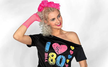 Eine Frau mit lockigem blondem Haar, einer rosa Schleife, einem rosa Netzhandschuh und einem schulterfreien T-Shirt mit der Aufschrift „I love the 80s“ posiert und lächelt vor einem schlichten weißen Hintergrund.